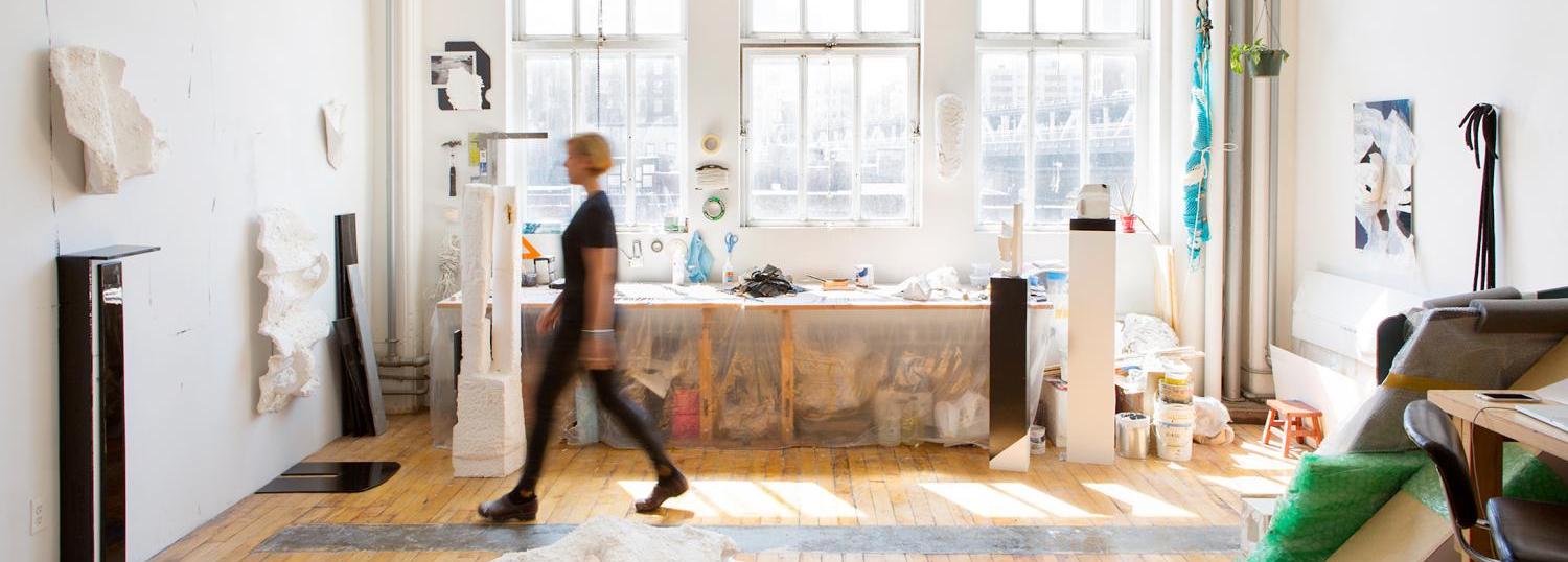 Person dressed in black walks across room with big windows across the back wall with lots of light. 