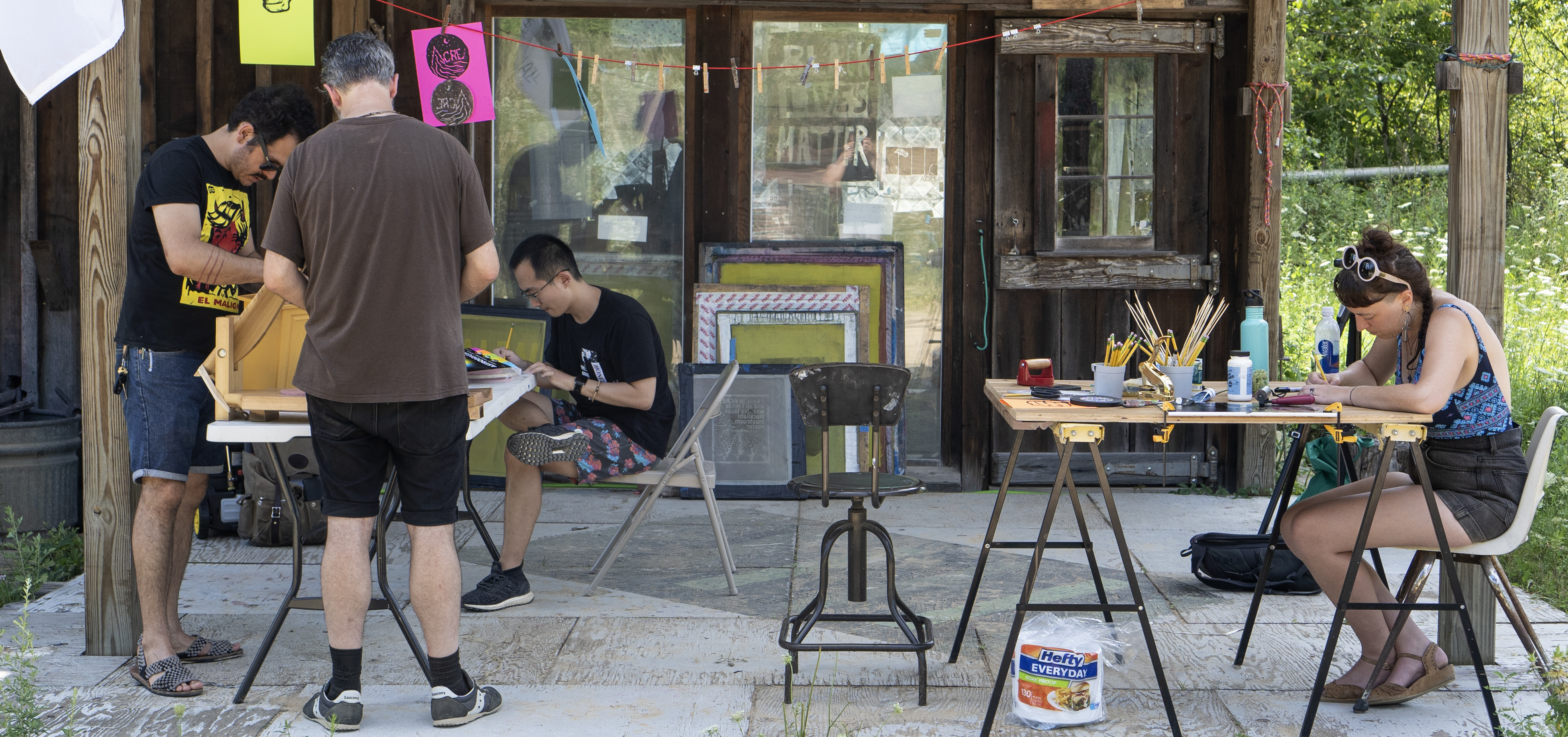 Four figures seated at tables with art supplies on a wooden outdoor porch, each focused intently on a different project.