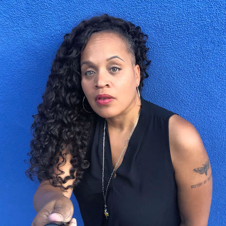 A photo of ACA CEO Lisa Funderburke with long black curly hair, a sleeveless black blouse standing in front of a blue wall