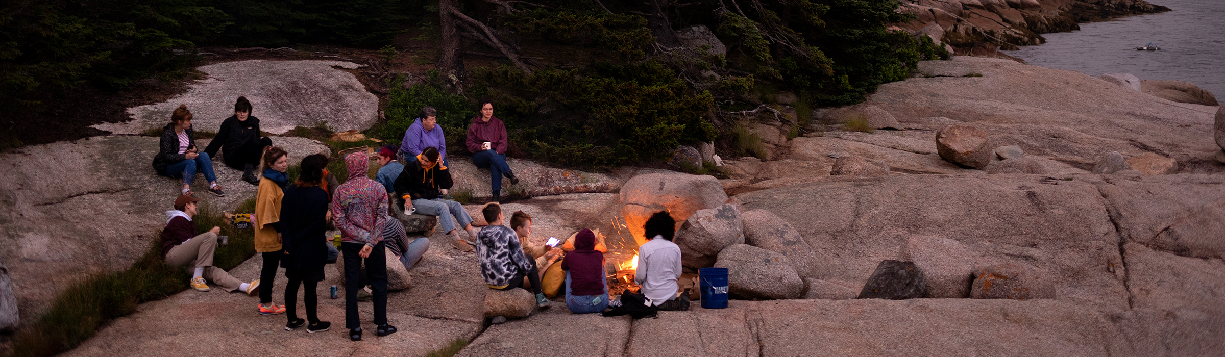 Artists gather around a campfire on a rocky shoreline