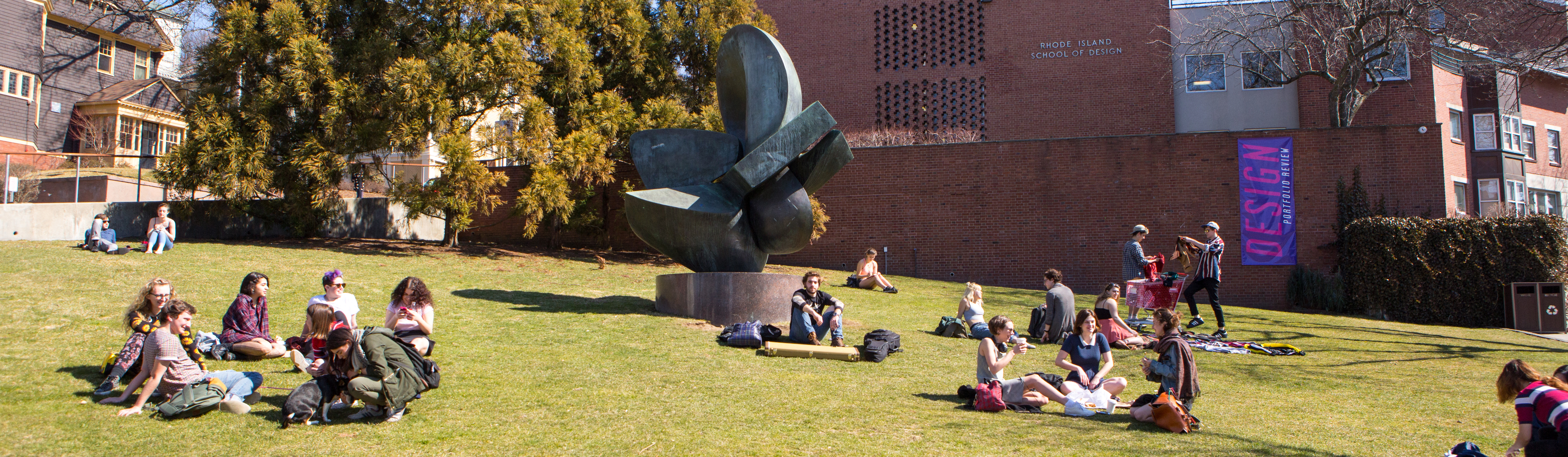 Groups of students relax together in clusters on a green lawn in front of a sculpture