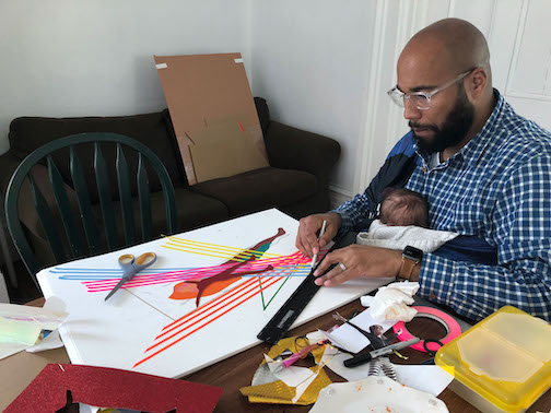 A person sitting at a table holding a pencil and ruler working on a piece of art. A baby is resting on their chest. 