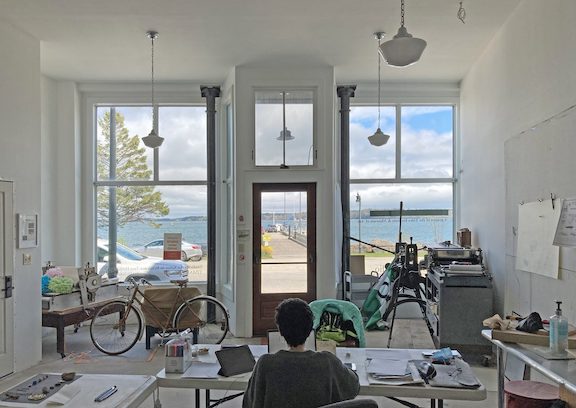 A person with their back facing the camera in a studio space with two large windows facing a body of water. 