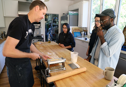 A chef demonstrates a pasta-making machine in action to three watchful artists.
