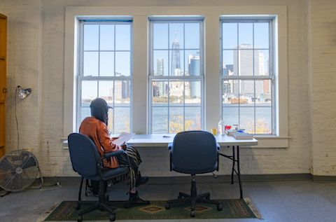Artist Asiya Wadud sitting at her desk facing the window at Governors Island overlooking the Manhattan skyline.