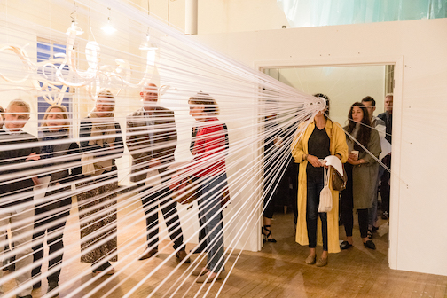 A group of people in a gallery space. One person is looking through an interactive art piece. 