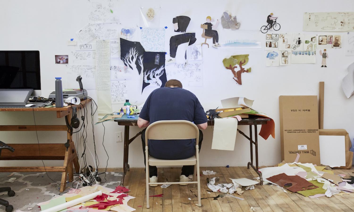 Back of artist at table with drawings on the wall. Photographer: Brad Ogbonna.