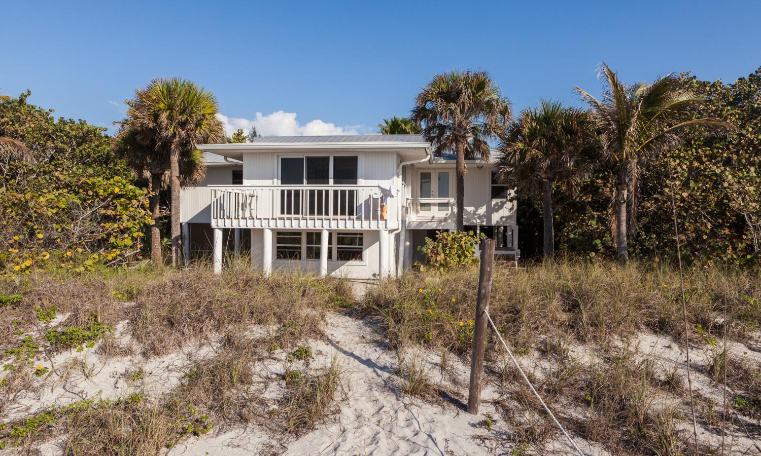 A white house surrounded by palm trees in the background with sandy dunes in the foreground