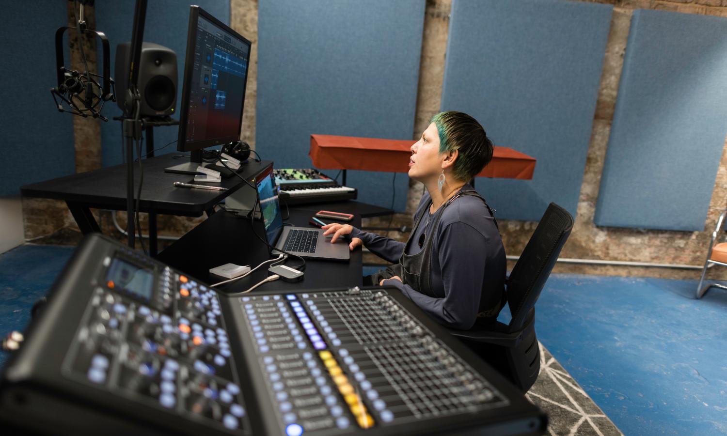 Carmina Escobar sits at a table in the Sound Studio with a computer, sound board, and monitor in front of them. Blue sound barriers line the brick walls behind her.