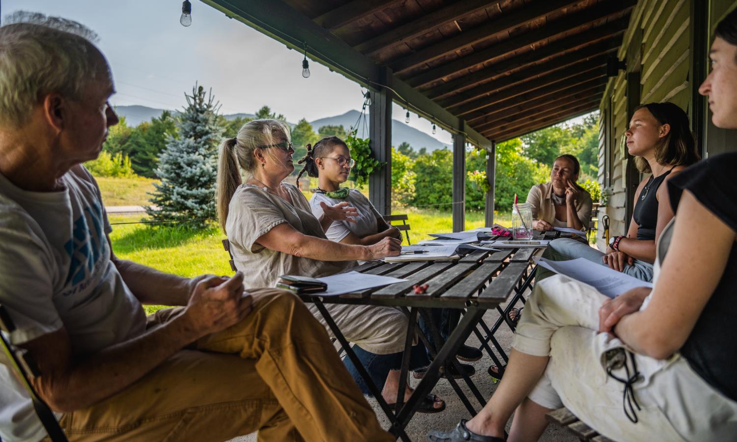 artists sitting on a porch around tables talking, with views of the mountains on a summer day
