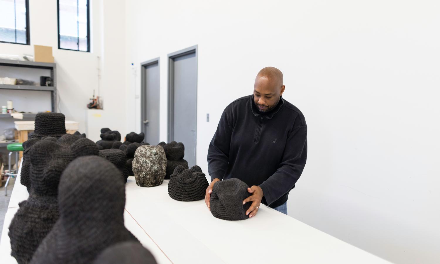Donté Hayes stands at a table in the Okada Sculpture + Ceramics Facility holding black, textured ceramic sculptures with several other sculptures surrounding them.