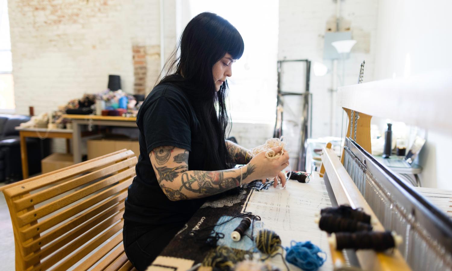 Erin M. Riley sits at a floor loom while weaving with several colors of yarn and string in their live/work studio.