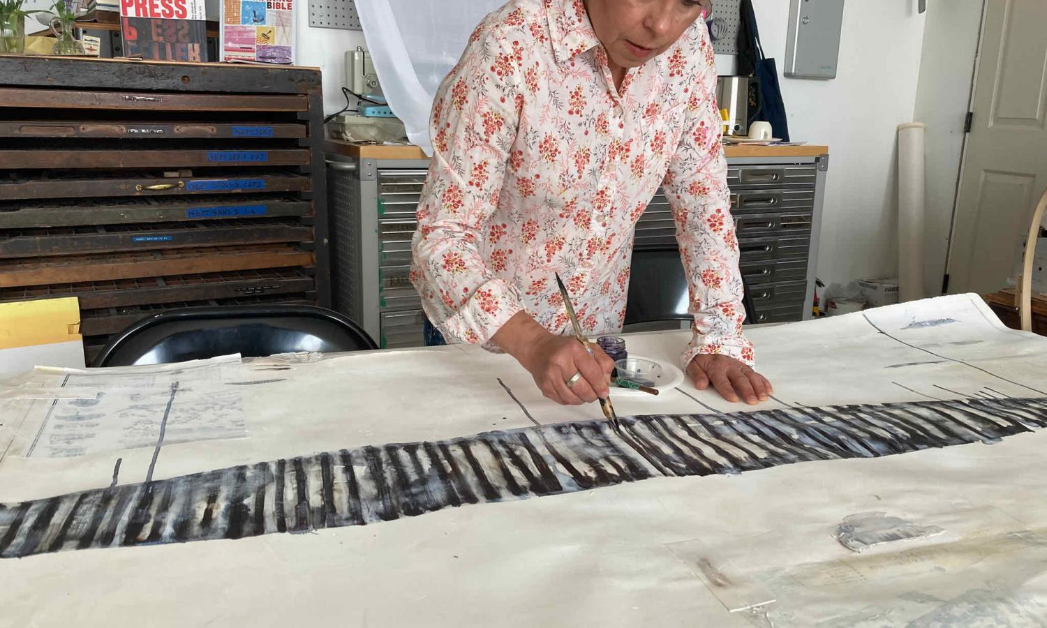 Luba Zygarewicz stands at a studio table, painting inked lines on paper and mixed media.