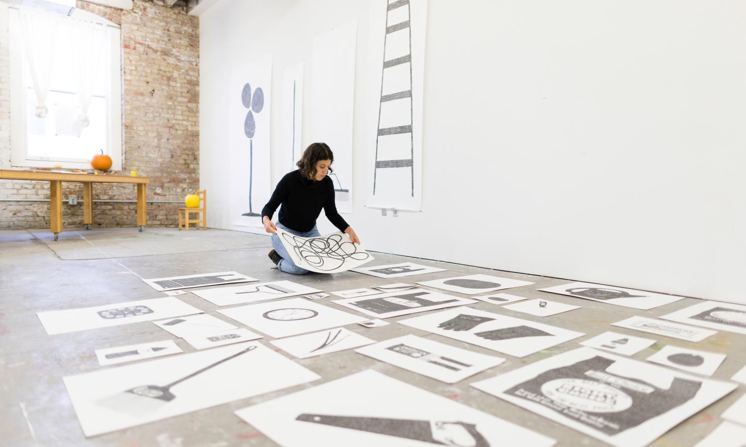 Mimi Laquidara kneels on the floor in front of a several black and white drawings arranged on the floor and on the wall of their live/work studio.