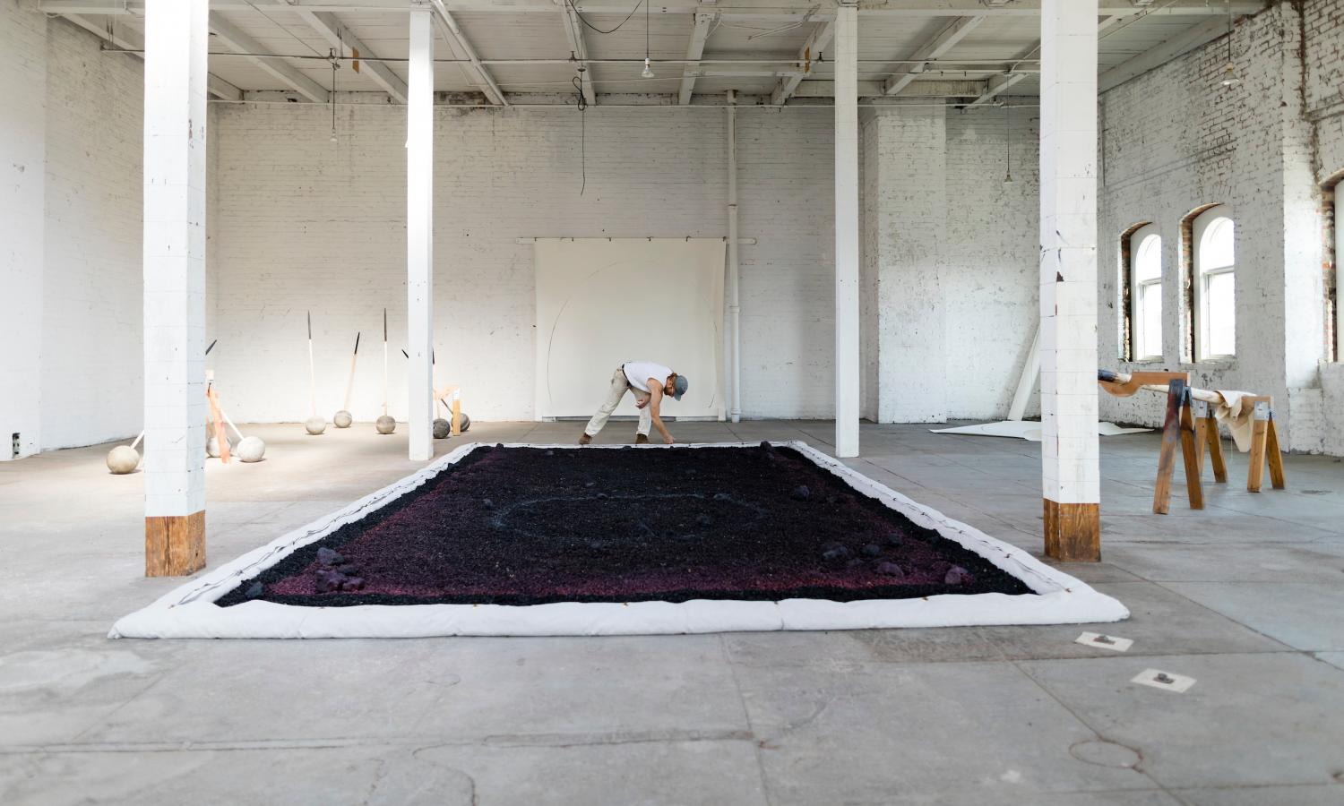 Nicolaus Chaffin arranges small rubber tire pieces to fill a large, rectangular shape onto the floor of 5th floor within several floor to ceiling columns.