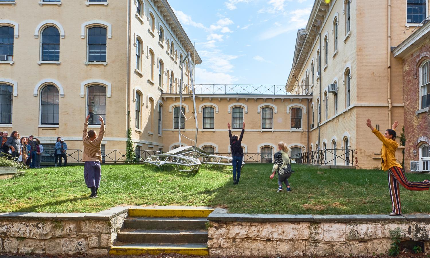 Members of Kinesis Project dance theatre move in various poses on the lawn in front of historical buildings and a large sculpture of a vessel by Tattfoo Tan