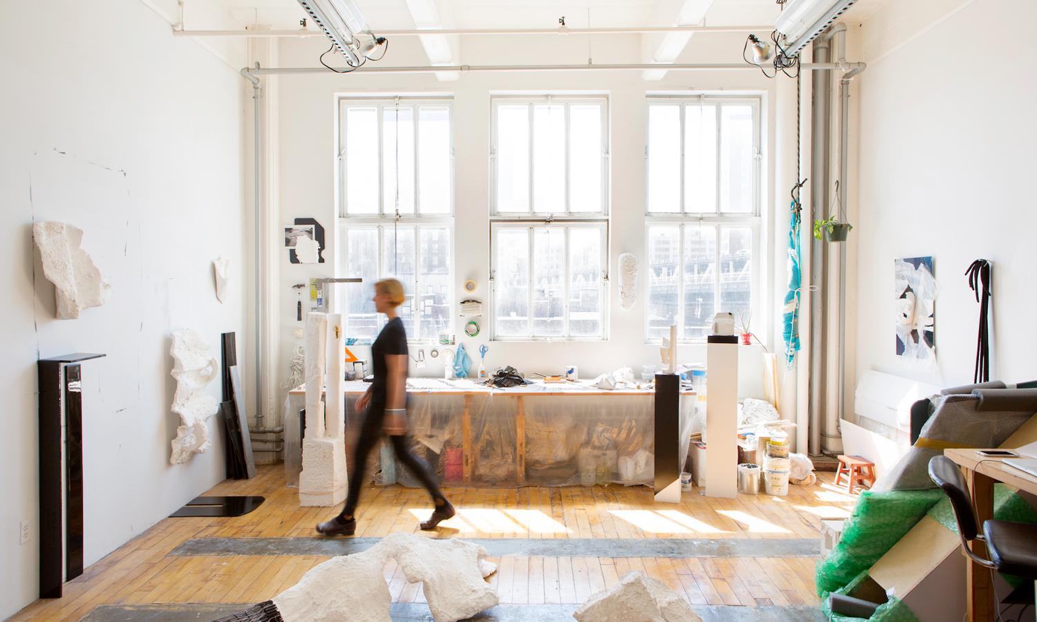Person dressed in black walks across room with big windows across the back wall with lots of light. Photographer: Nina Choi