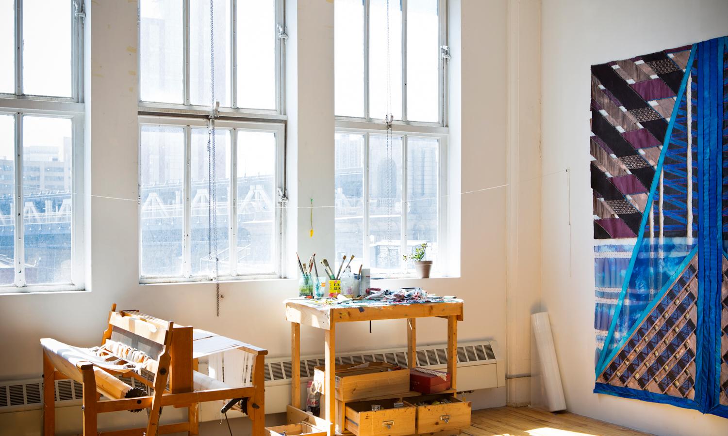 A wood loom is in front of large windows with bright light streaming through and a blue fiber artwork on the wall to the right. Photographer: Nina Choi