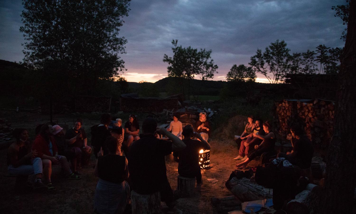 A large group of people gathered at dusk sitting on tree stumps and log benches around a glowing fire pit.