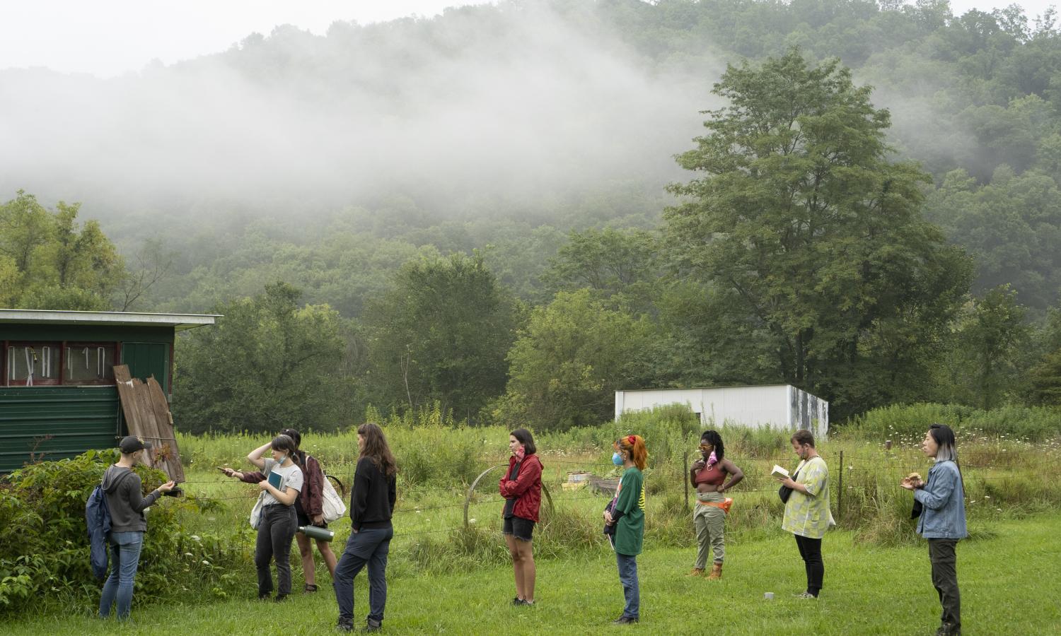 An attentive group faces a speaker in front of a foggy lush green backdrop at the bottom of a sloping hill.