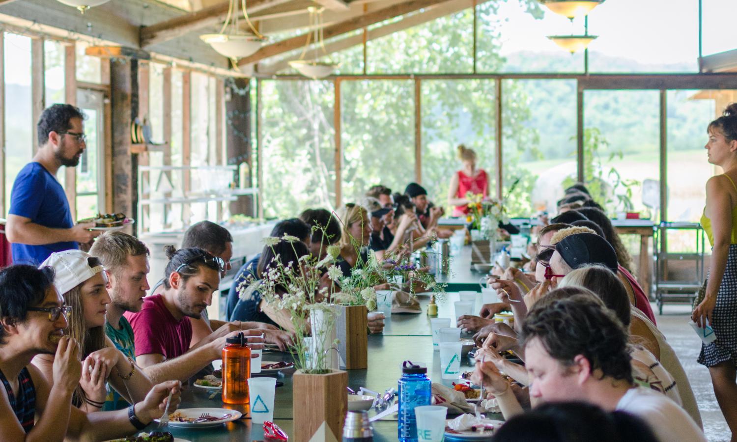 View from the head of a long shared table in a screened-in kitchen flooded with daylight where several people facing one another eat and talk.