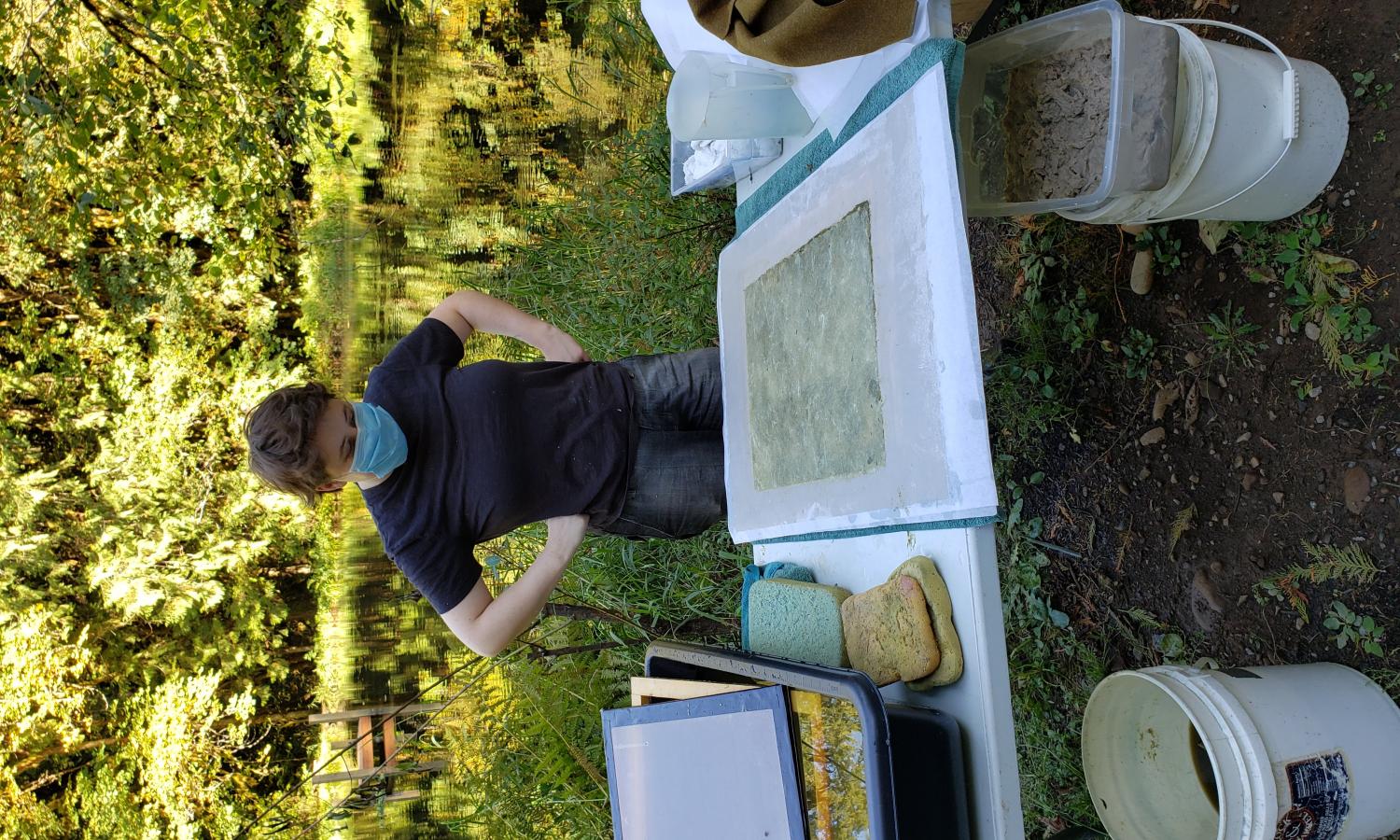 Paper resident Maddy Dubin looks at a sheet of handmade paper they just made in front of a green forested background with pond.