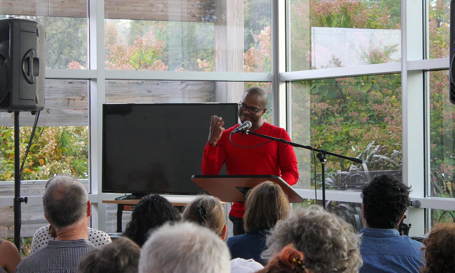 Resident in red shirt speaking to seated audience in front of glass windows with visible sound system and tv monitor