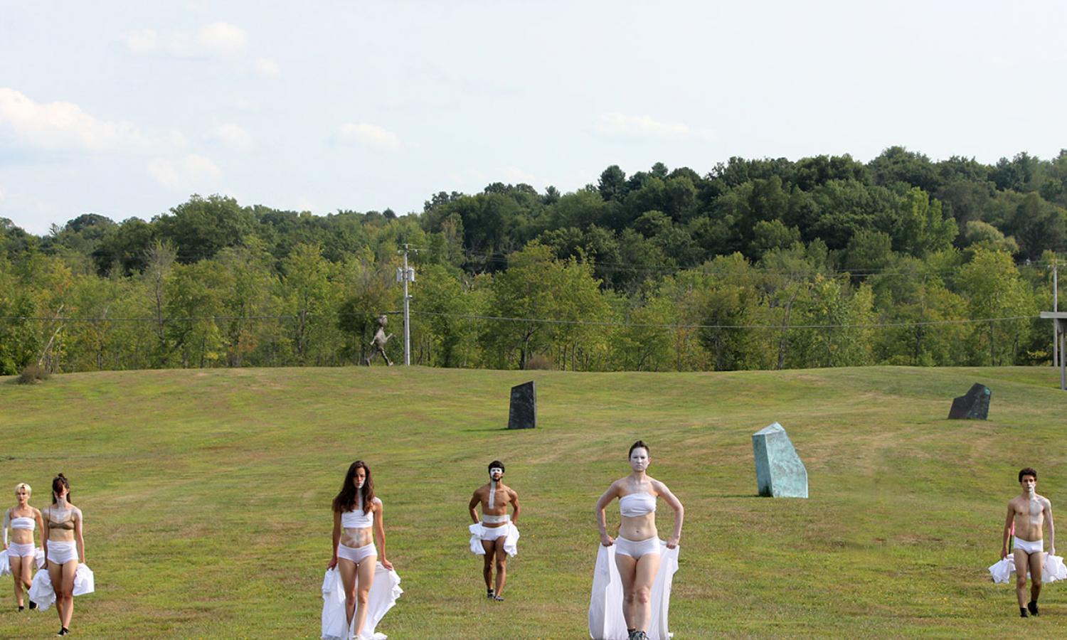 Performers in white outfits and face paint in outdoor landscape with sculptural elements
