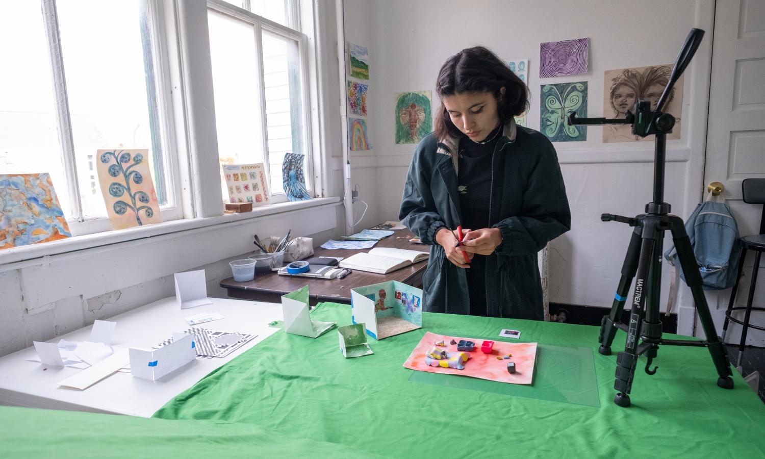 Photo of Laura Medina working at table with green screen cloth. Laura is working on a stop motion paper project.