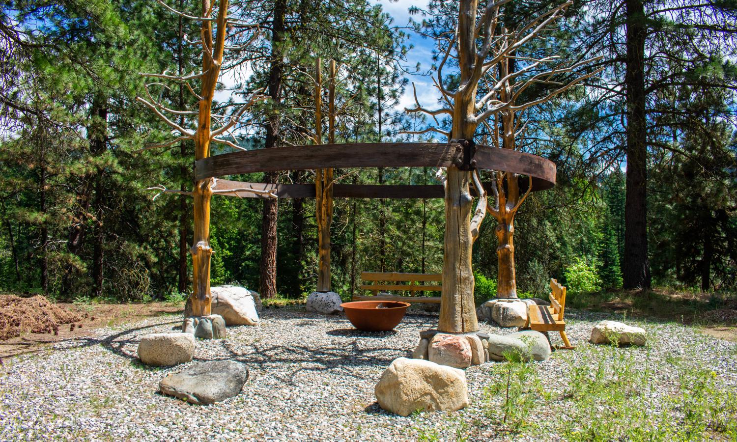 Finisterre: Four peeled, stained, and weathered ponderosa pines standing in a circle bound by an iron band connecting them. Larger rocks surround the trees representing artists who have been influential and important to the community of the Guild.  Background is ponderosa pines and foreground is a pathway of smaller rocks leading up to the piece.