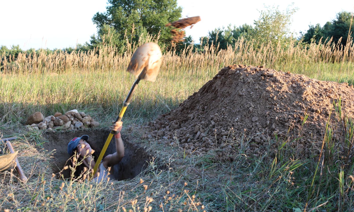 A person is pictured shoveling dirt from a grave that is being dug for an installation. Golden light catches the background and the shovel in the air. They are deep enough that only their head and forearms can be seen as they sling the dirt to the top of a dirt pile to the right of the image. The are shoveling with a round point shovel with a yellow and orange handle. The arist is white. The are wearing orange gloves, a white tank top, and a blue hat.