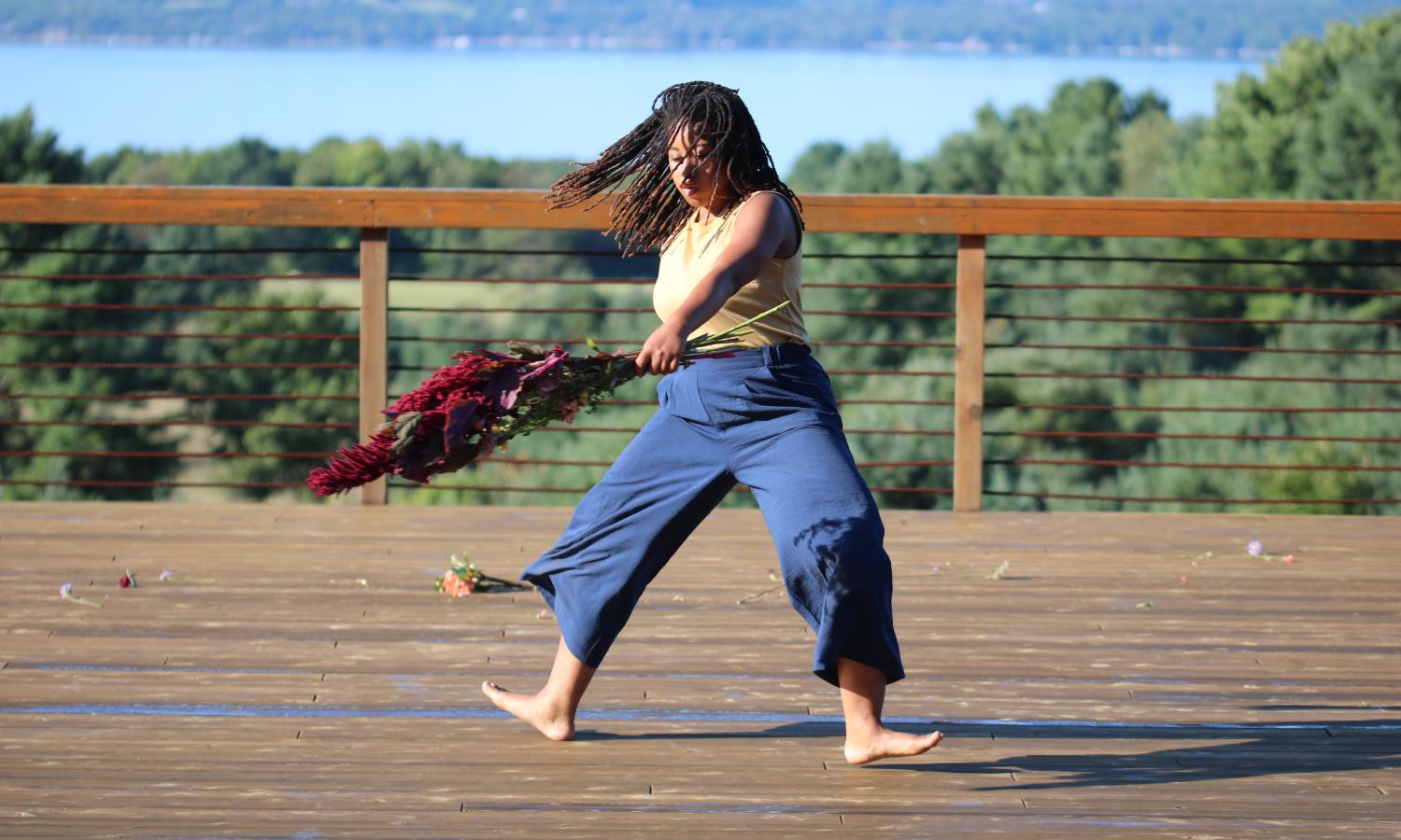 A dancer can be seen in the center of the image on a deck. They are black with long braides. They are wearing a mustard colored tank and navy pants. They are caught in a moment of spinning, holding flowers in each hand. Their left arm is in front of them and the right arm is slightly behind their body. Their feet and legs are fully turned out with the weight caught in this moment on both heels. The background of the image is trees, a field, and a lake.