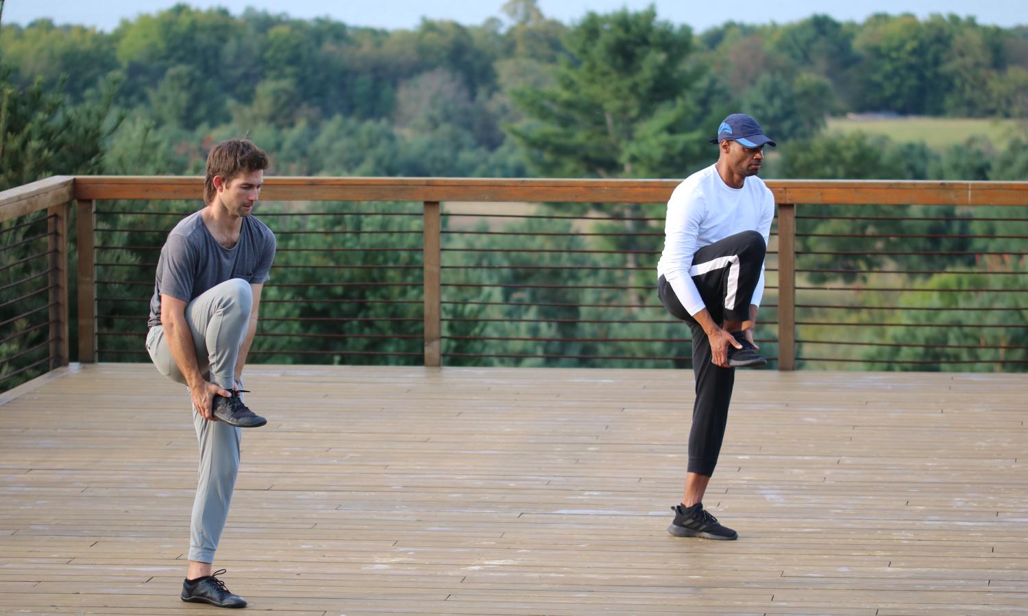 Two dancers are on a cedar deck. In the background, out of focus, are trees, fields, and a lake. The dancers are in the same pose. Perched on their left foot standing upright. They are holding the bottom of their right foot with both hands with their gaze ahead of them. The dancer on the left is white. They have brown hair and are dressed in gray. The dancer on the right is black. They are wearing a blue cap, white shirt, and black pants with a white stripe.