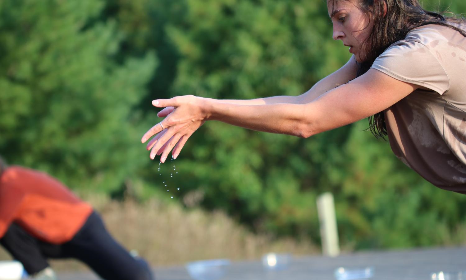 Two dancers are on a cedar deck. One dancer on the right is in focus. Only their torso is visible. They have their arms outstretched, water dripping from their hands. They have dark hair that is wet, and are wearing a tan top. The deck and the bowls ful of water are out of focus. The image background is white pines. The second dancer is in a deep squat with their torso reaching out of the image. They are in an orange top and black pants.