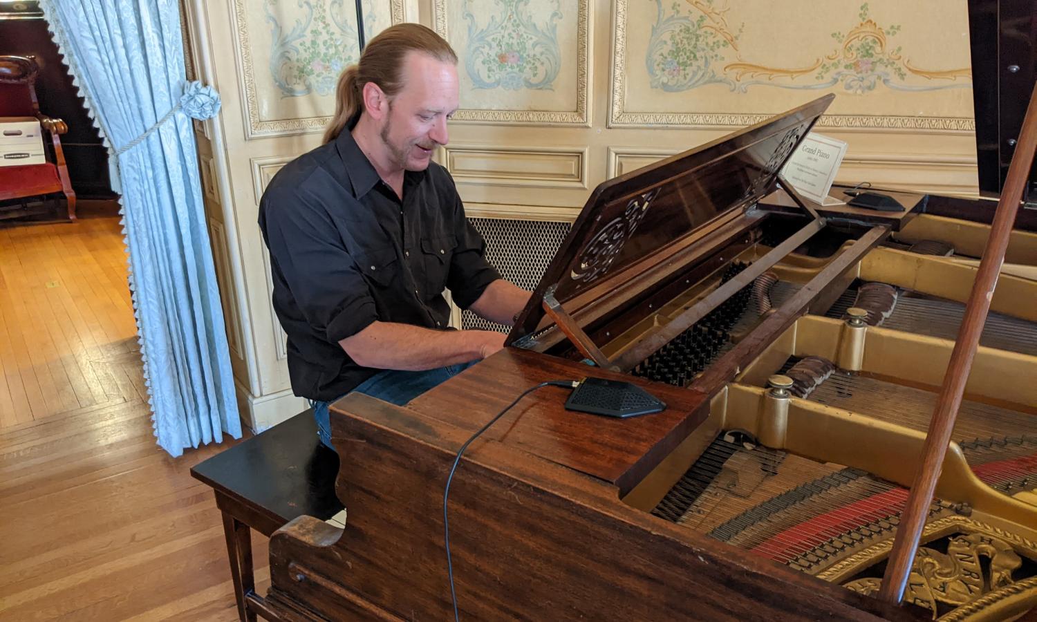 Artist-in-Residence Aaron Jennings sits at an elaborate but aged grand piano. He is smiling while he plays the piano. He is sitting in lavish historical room in the Historic Clark Chateau in Butte, Montana. The walls are cream colored with elaborate paintings and moldings.