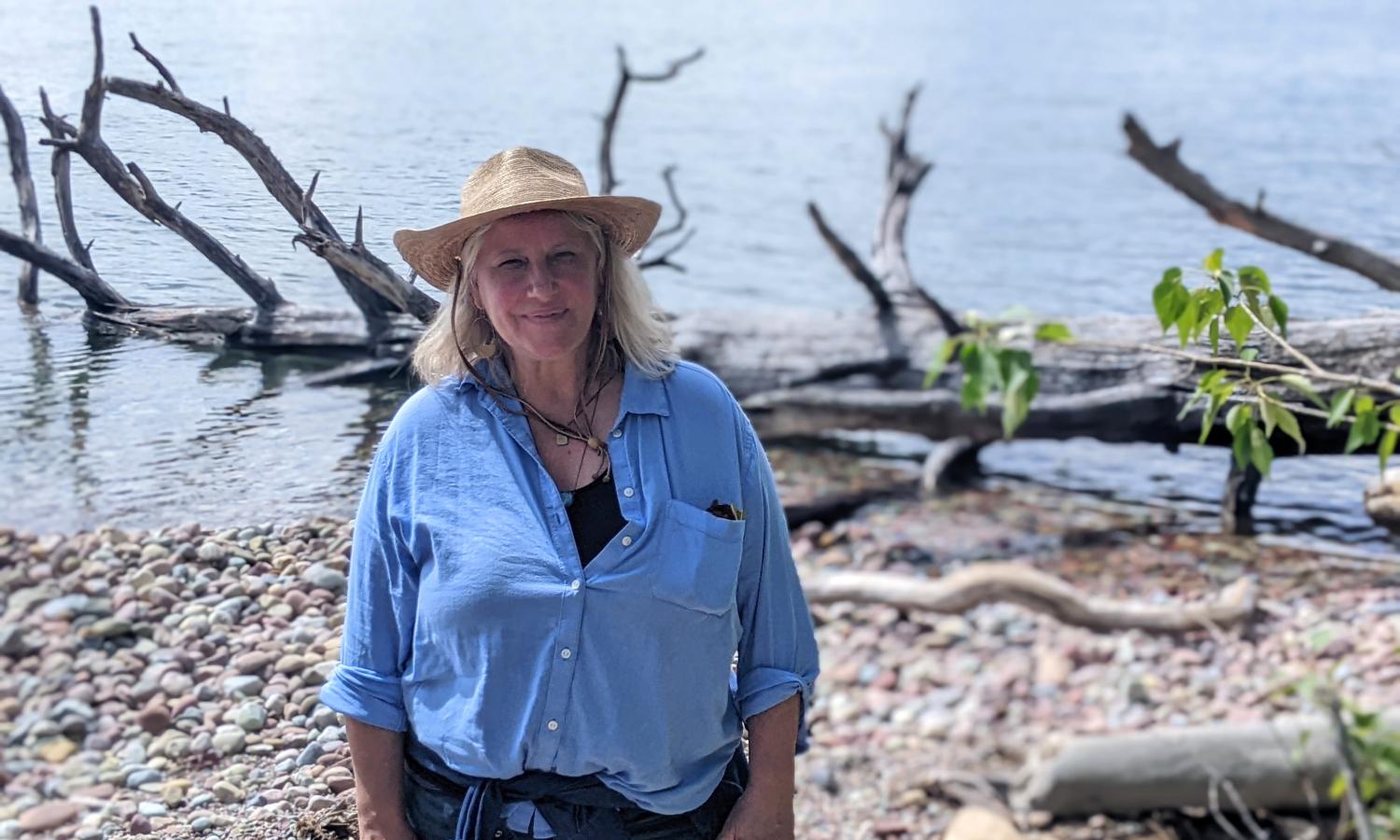 Artist Jennifer Ogden stands in front of Flathead Lake in Polson, Montana. She is smiling in a wide brimmed hat in front of the beautiful blue summertime water.