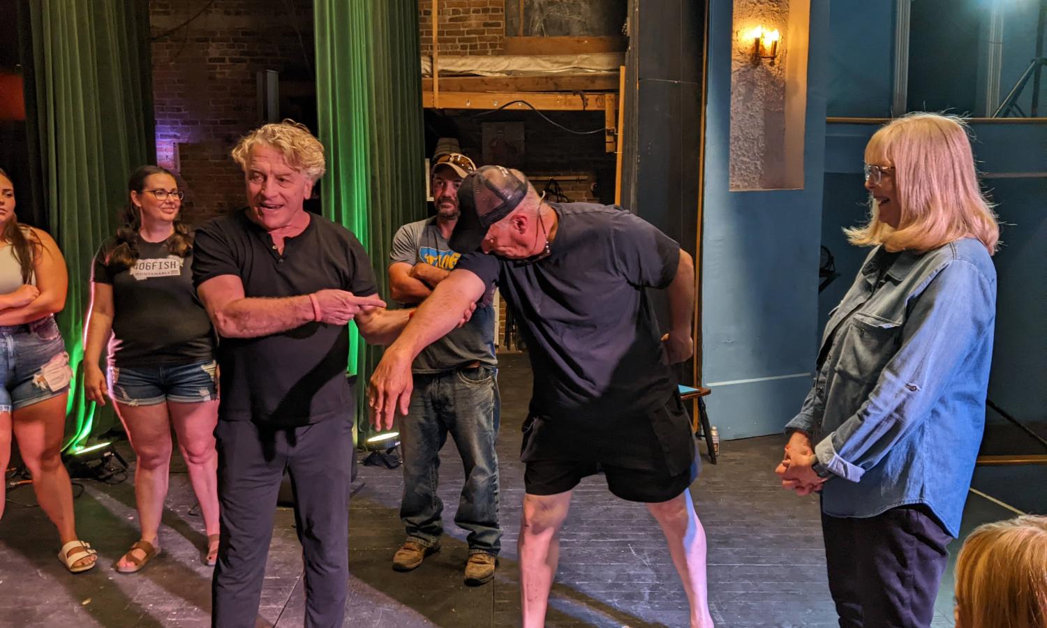Actor Bill Bowers is standing on a stage in a historic theater. He is teaching a movement workshop to a group of people of all ages. Bill is holding the arm of one of the participants, while the participant holds his arm to his face, like the trunk of an elephant.