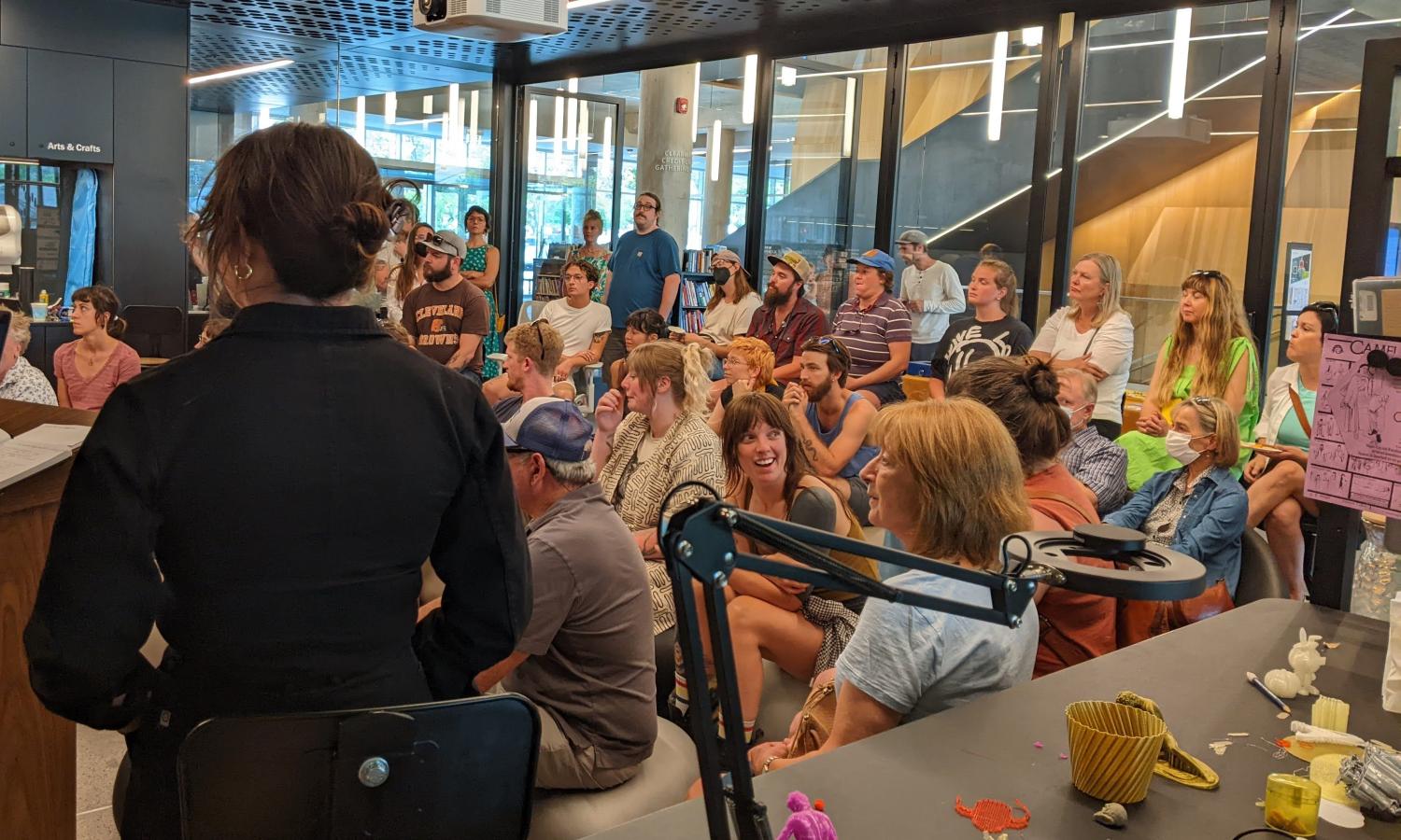 We see the back of Artist-in-Residence Cait Finley, who is giving a presentation to a crowd of 30 people sitting close to each other in a glass walled room -the MakerSpace of the Missoula Public Library.