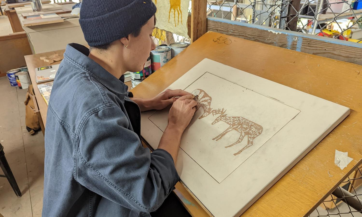 Artist-in-Resident Ian Hanesworth sits at a drafting table located in a mezzanine, surrounded by paint cans and art supplies. Ian is using beautiful Japanese woodcarving tools to carefully carve an image of two deer facing each other.