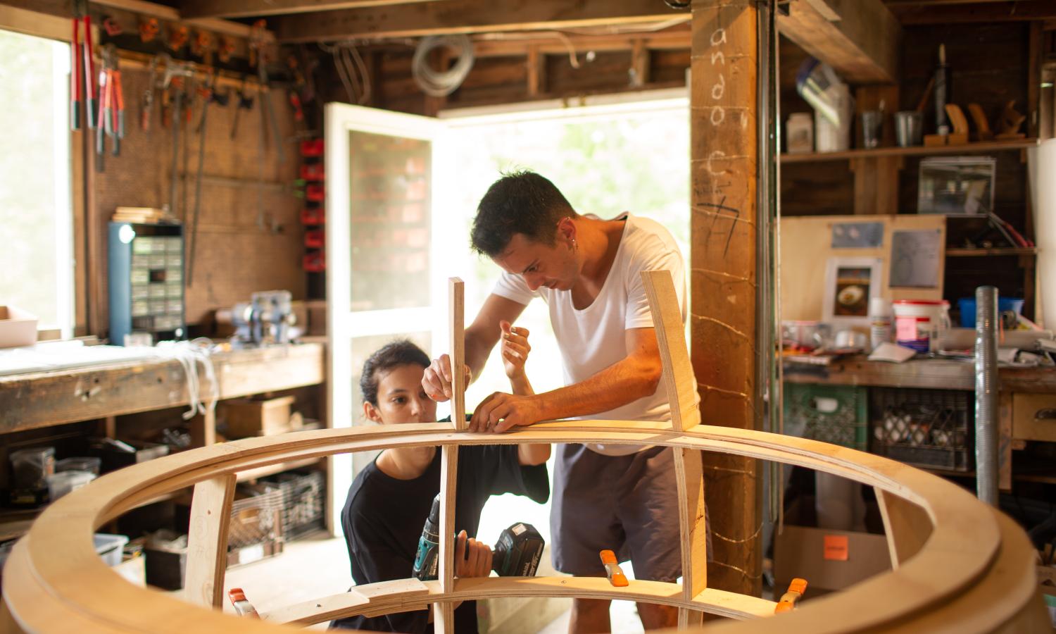Two individuals build a large, rotund wooden structure in a woodshop.