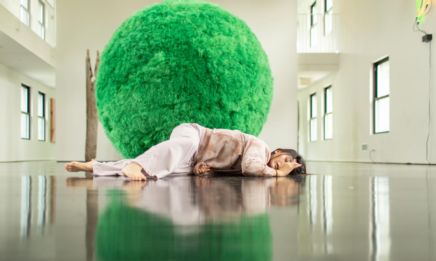 A performer dances on a wooden floor in front of an over-sized ball covered in green fur.