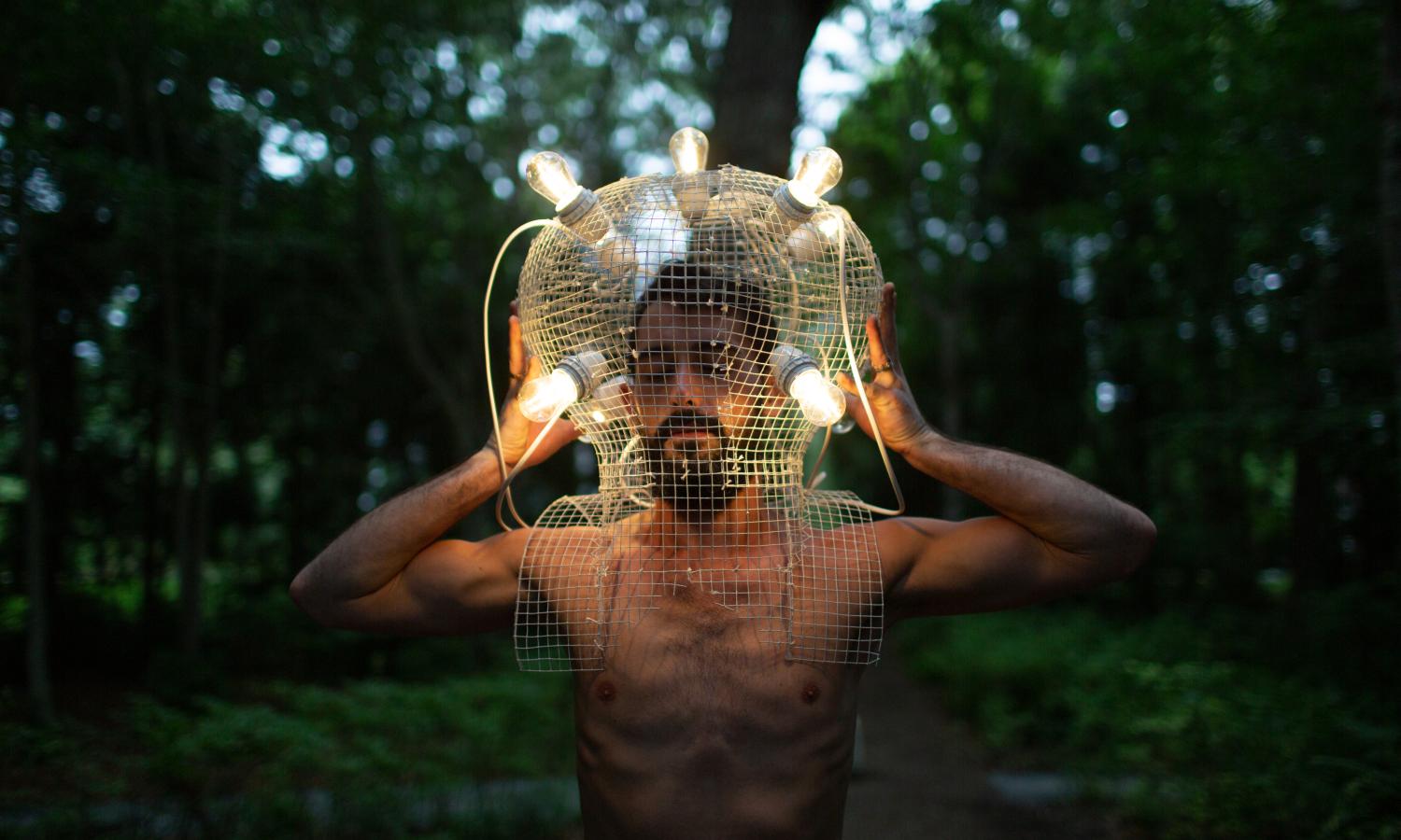 A bare-chested performer holds a large white-wire mesh helmet on his head, with lit lightbulbs scattered around his head, providing light against the dark, forested backdrop.