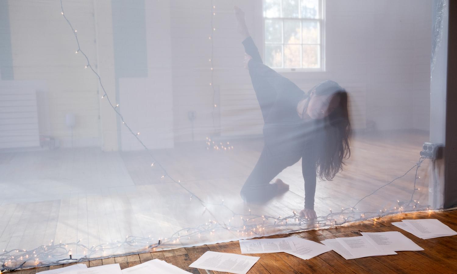 Shot of a room with a thin translucent white sheet in front of dancer Ari Mokdad who is posing with one hand and one knee on the floor, paper with text is scattered about the rest of the floor.