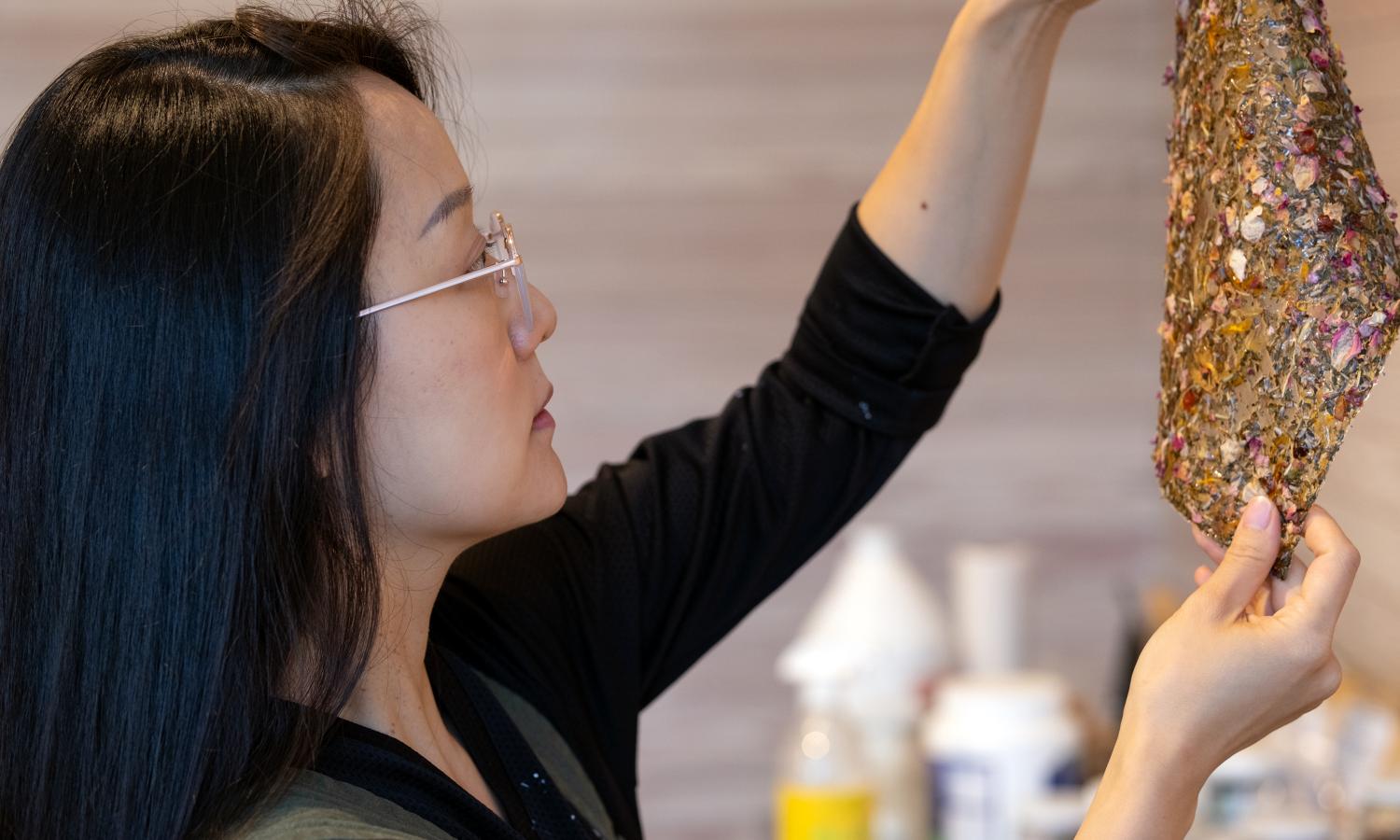 Josephine Lee in profile, holds up a translucent bio-material substance with plant materials embedded in it.