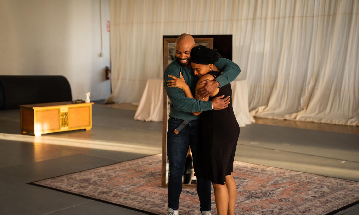 Two Black people are hugging. They are standing on an ornamental rug and the back wall is covered in a flowing curtain-like material.