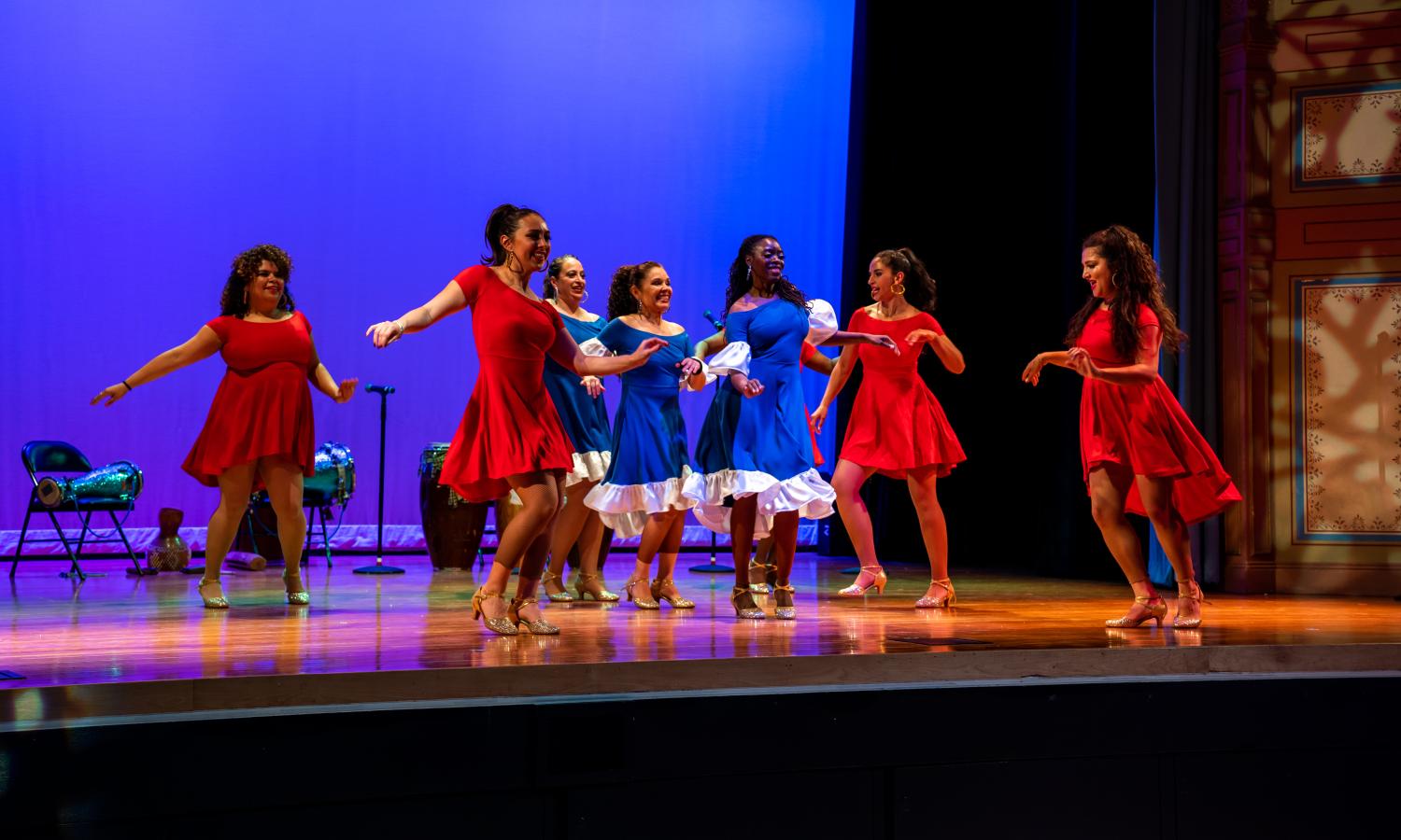 A group of people are dancing on stage in two concentric circles. The inner circle is wearing blue Afro-cuban attire and the outer group is wearing red Afro-cuban attire. The backdrop of the stage is colored with blue lighting.