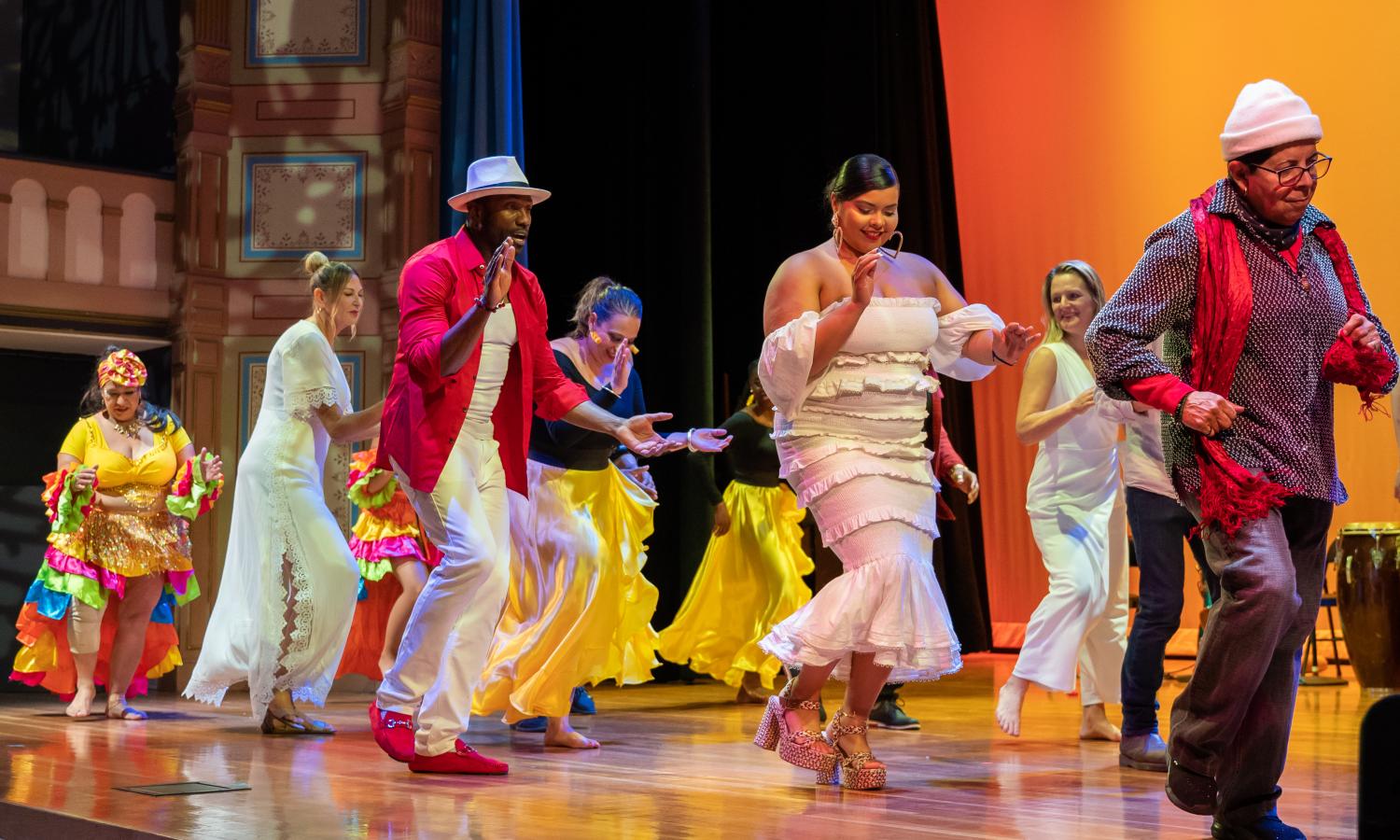 A group of people are standing on a stage wearing colorful, traditional Afro-cuban clothing. They are dancing and moving to the right of the stage. The backdrop of the stage is colored with orange and yellow lighting.