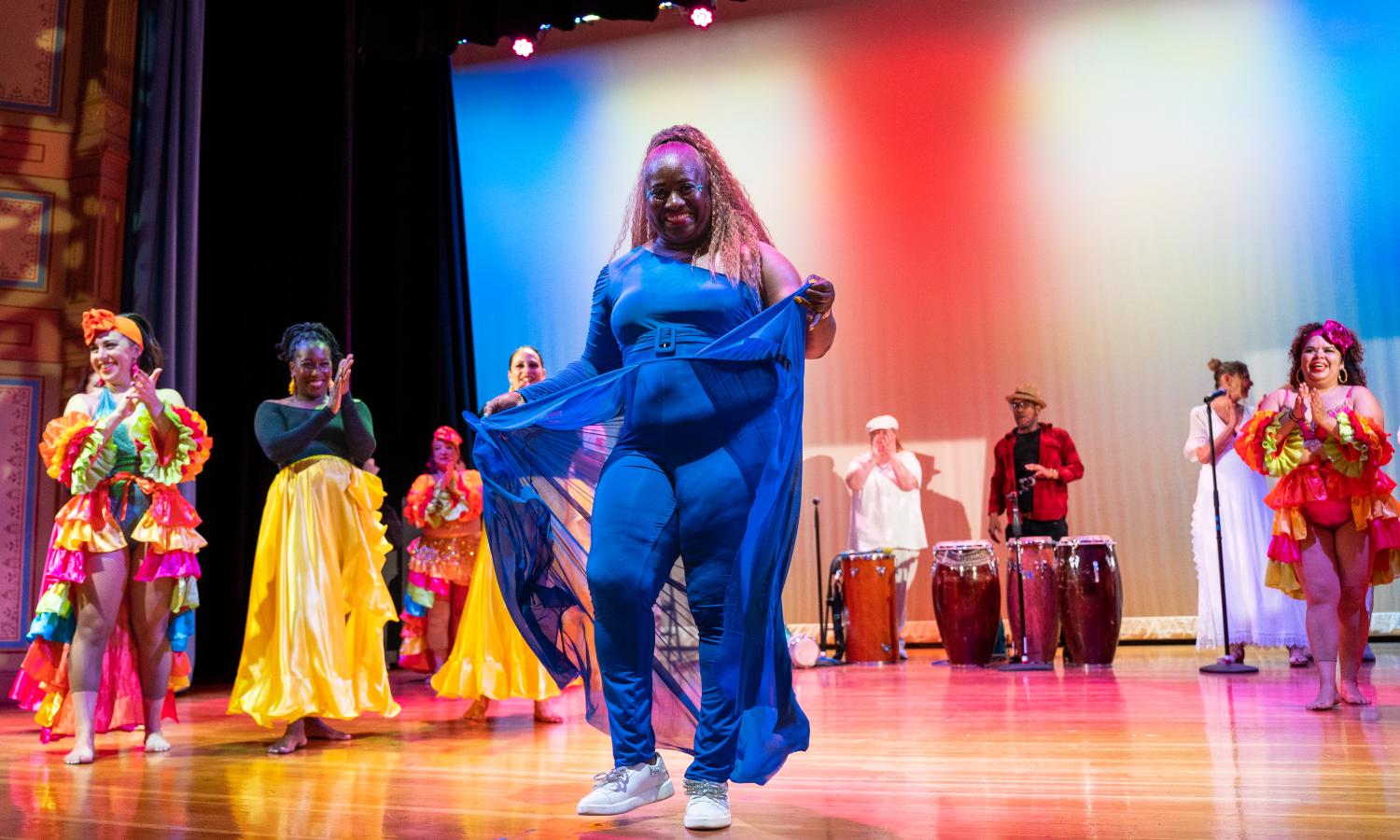 A group of people are standing on a stage wearing colorful, traditional Afro-cuban clothing. There is a woman in a blue outfit dancing in the center. The backdrop of the stage is colored with red and blue lighting.