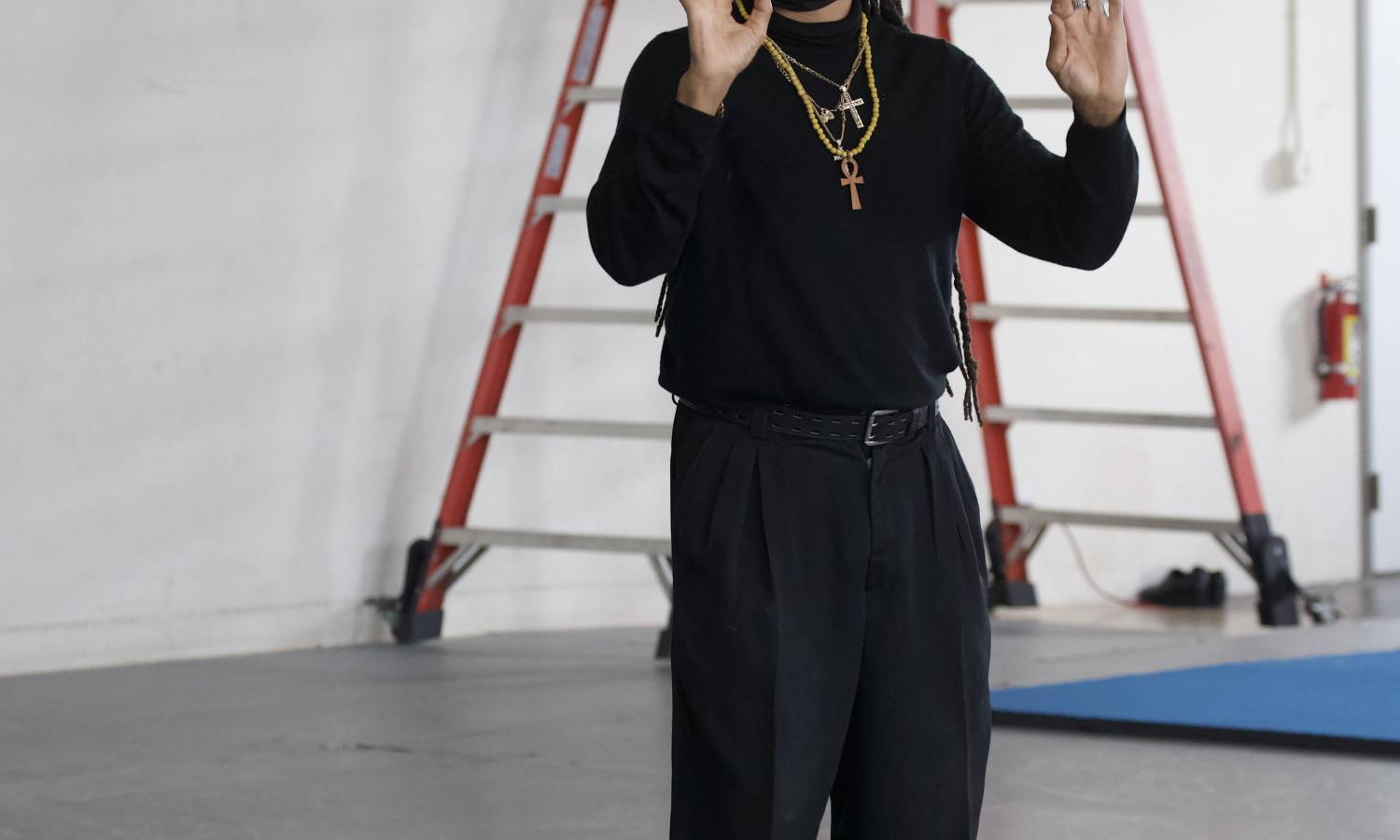 Black person wearing black stands with hands about face level. They are communicating using ASL