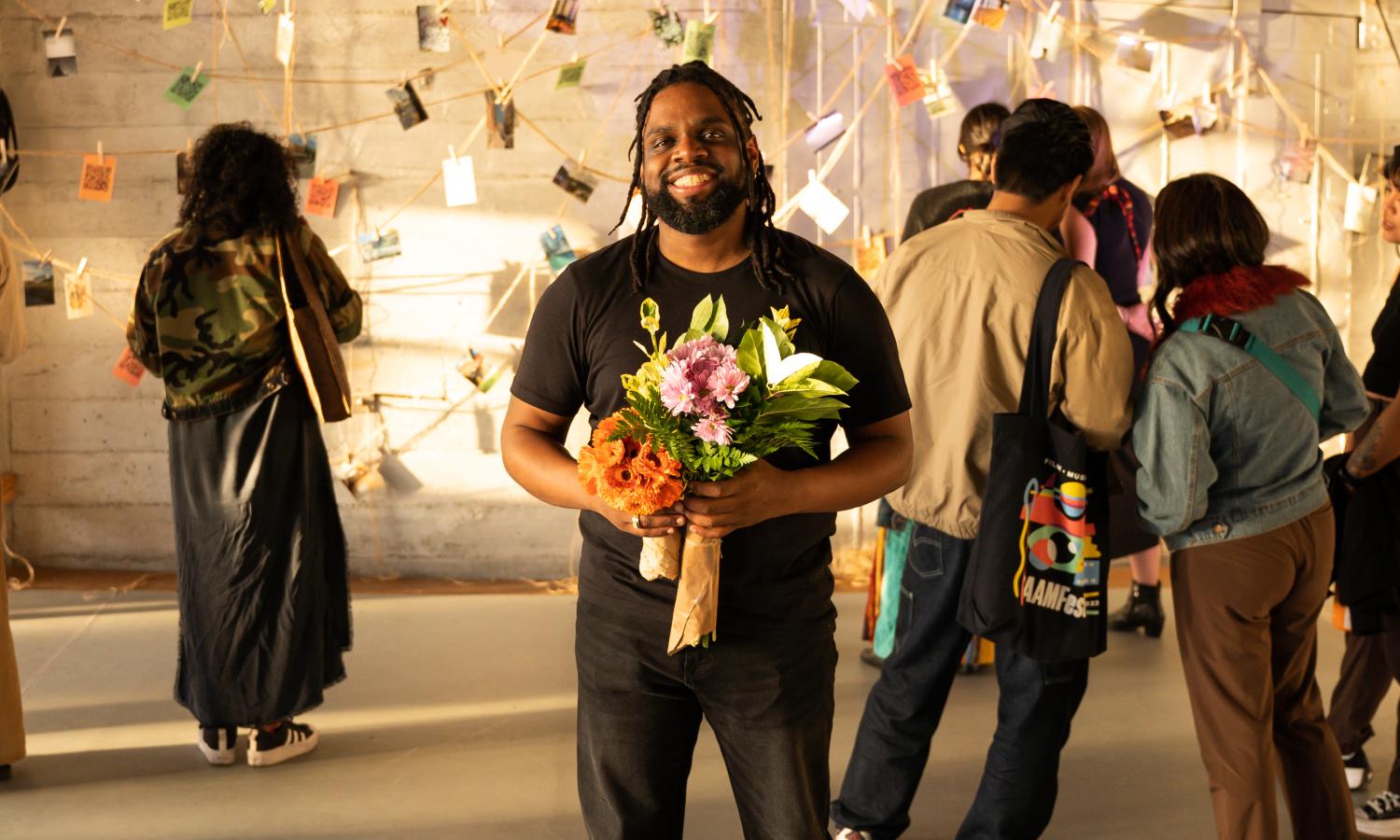 In a crowd of people, a Black non-binary person with locs stands with flowers in his hands. He is smiling.